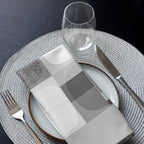 Folded grey cloth napkin featuring an abstract geometric pattern of circles and squares. Displayed on a plate with cutlery and a glass.