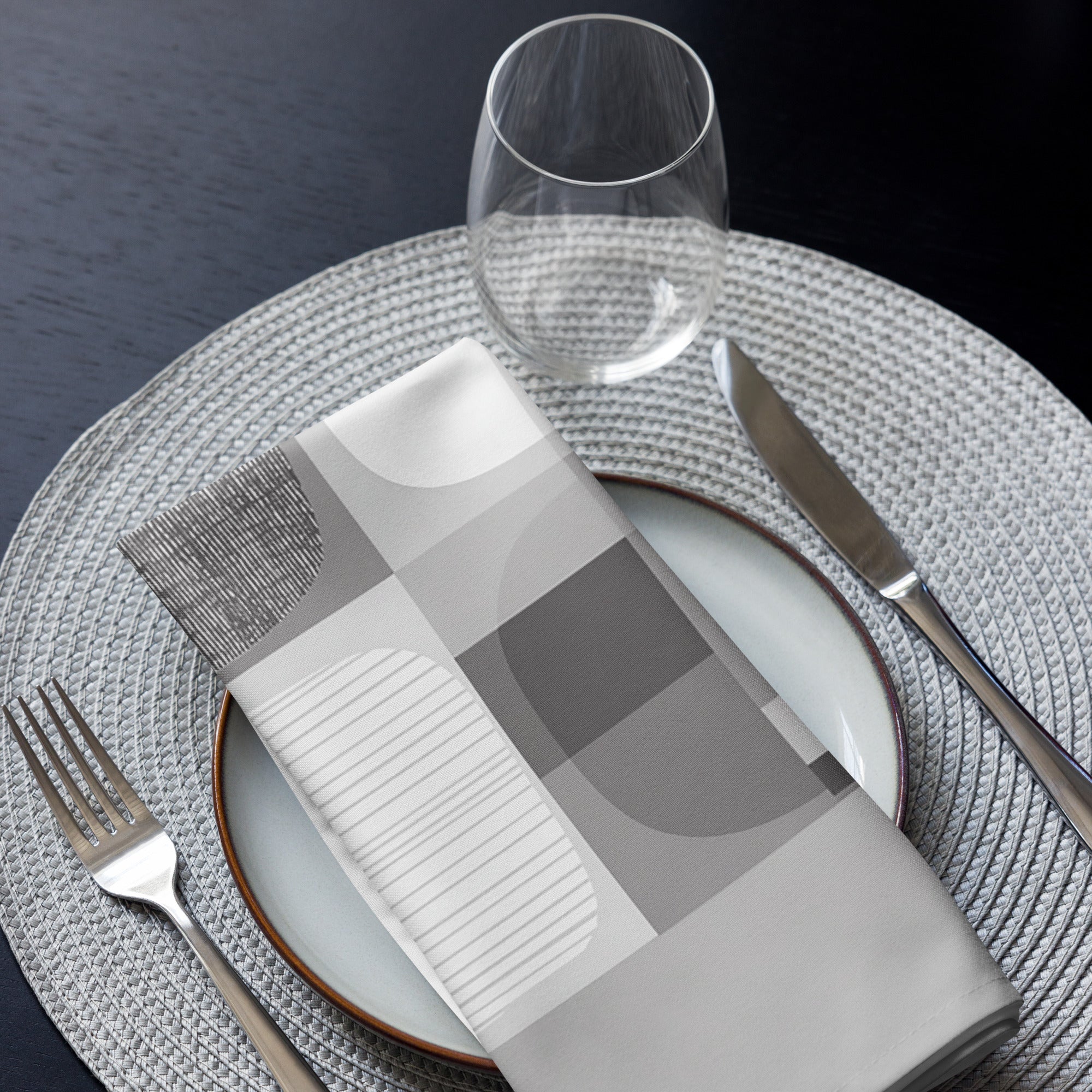 Folded grey cloth napkin featuring an abstract geometric pattern of circles and squares. Displayed on a plate with cutlery and a glass.
