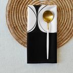 Folded black cloth napkin with an abstract pattern of eight white circles arranged in two rows. It is shown styled on a table mat with a gold and white spoon.