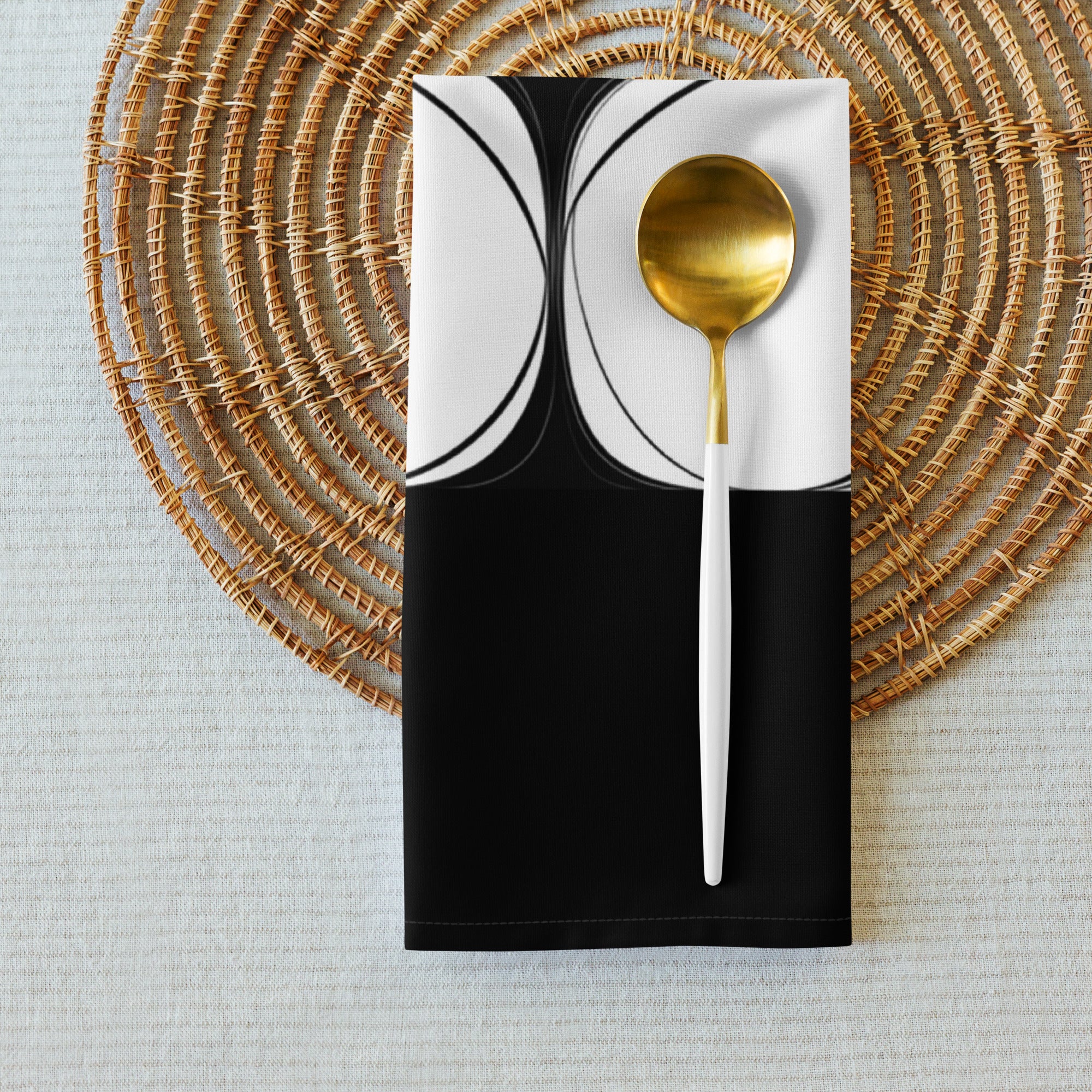 Folded black cloth napkin with an abstract pattern of eight white circles arranged in two rows. It is shown styled on a table mat with a gold and white spoon.