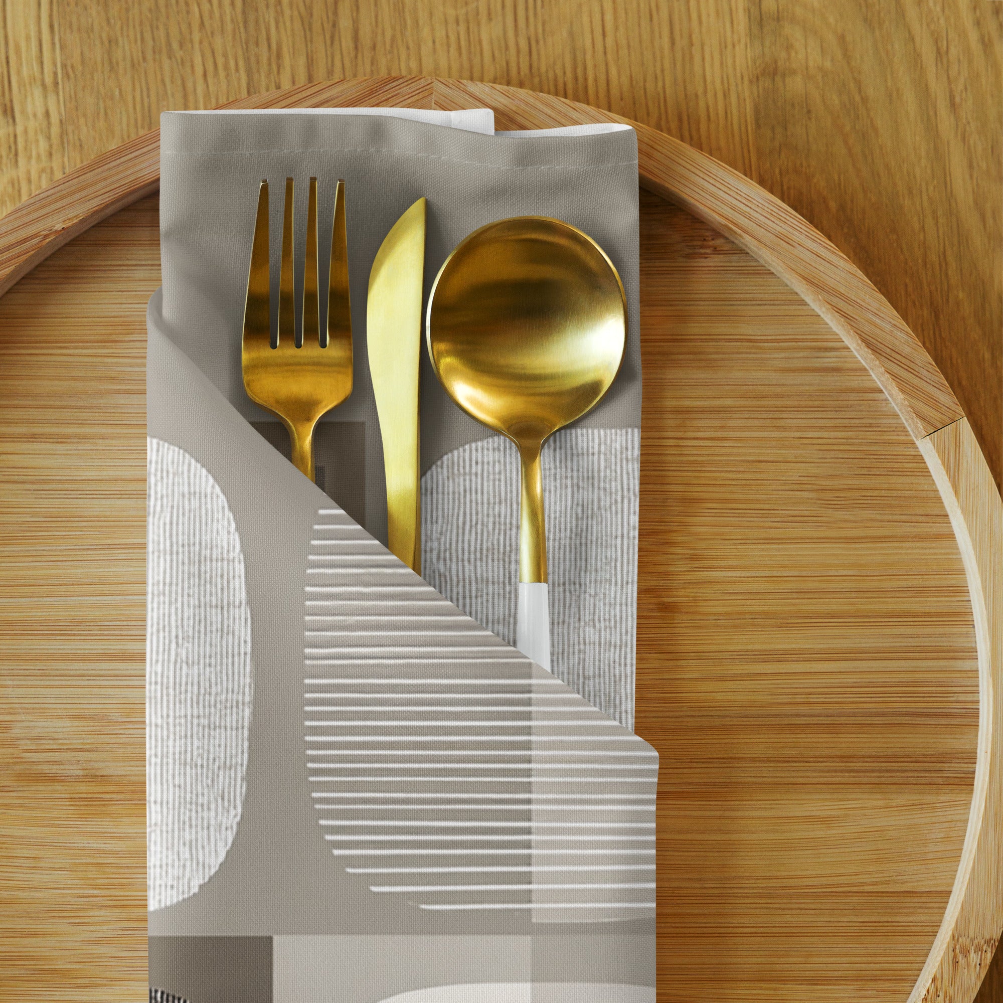 Sand-colored cloth napkin with abstract circles and squares, shown wrapped around cutlery on a wooden plate.